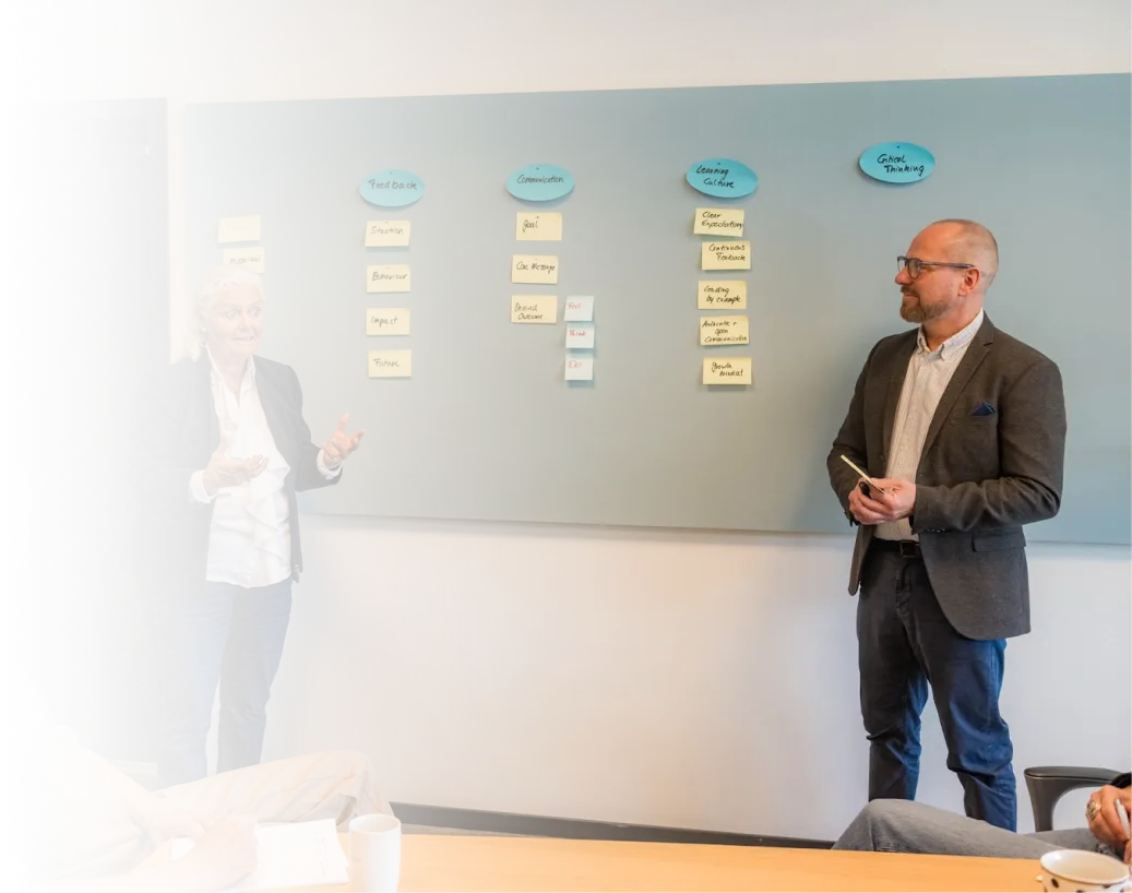 Two colleagues standing in a meeting room, facing each other while discussing with a grey board behind them displaying sticky notes organized under headings: Feedback, Communication, Learning Culture, and Critical Thinking.