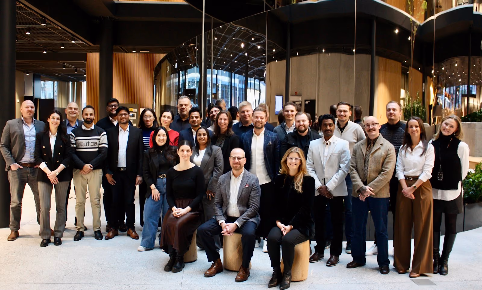 Group photo of 27 diverse professionals posing indoors in business casual attire, with some sitting on wooden stools and others standing behind, in a modern office lobby with reflective glass walls and plants.
