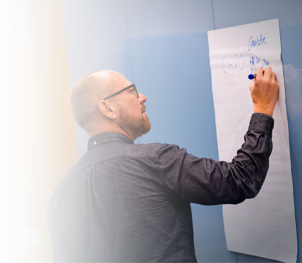 Man with glasses and a short beard writing on a large sheet of white paper mounted on a blue wall, using a blue marker.