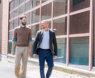 Two men walking outdoors beside a building with large windows, engaged in conversation and smiling.