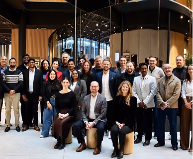 Group photo of 24 diverse professionals standing and sitting in an indoor modern office space with glass partitions and wooden panels, all facing the camera and smiling.