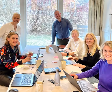 Six people gathered around a table in a bright room with large windows; five women seated with laptops and drinks, one man standing and smiling, suggesting a collaborative and friendly meeting.