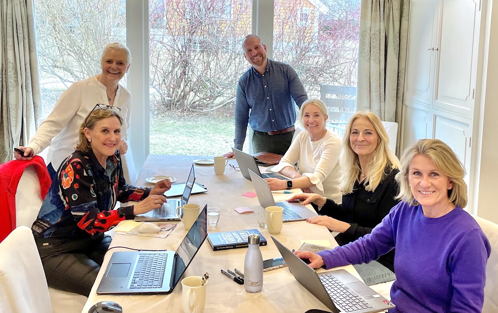 Six smiling adults, five women and one man, gathered around a table with laptops, notebooks, coffee mugs, and pens in a bright room with large windows showing trees outside.