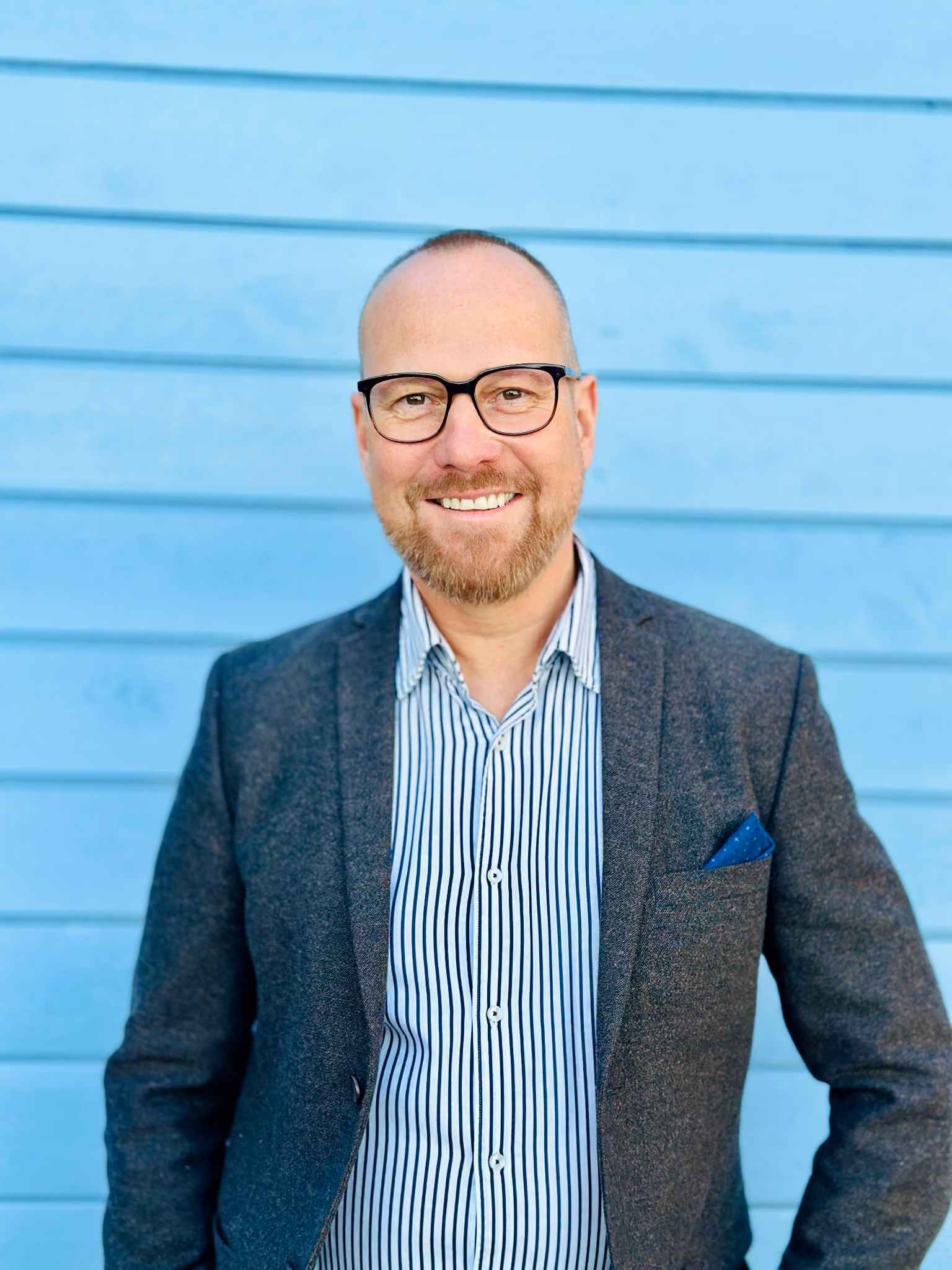 Smiling man with a beard and glasses wearing a striped shirt and dark blazer standing in front of a blue wooden wall.