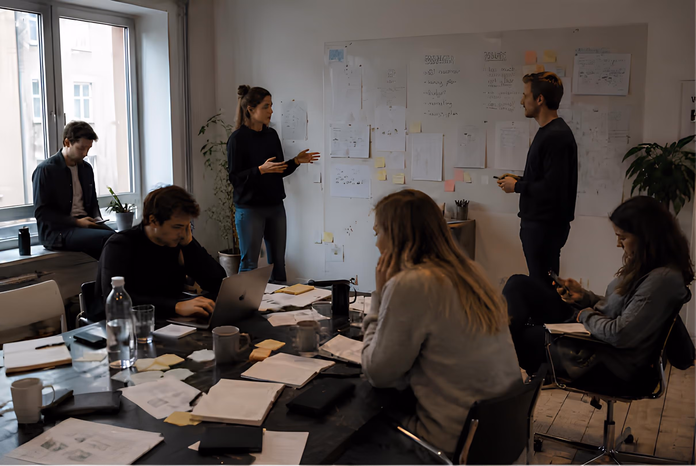 A group of six people in a meeting room with large windows; two stand near a whiteboard covered with papers and sticky notes while others sit around a cluttered table with laptops, notebooks, and drinks, some looking at devices or working.
