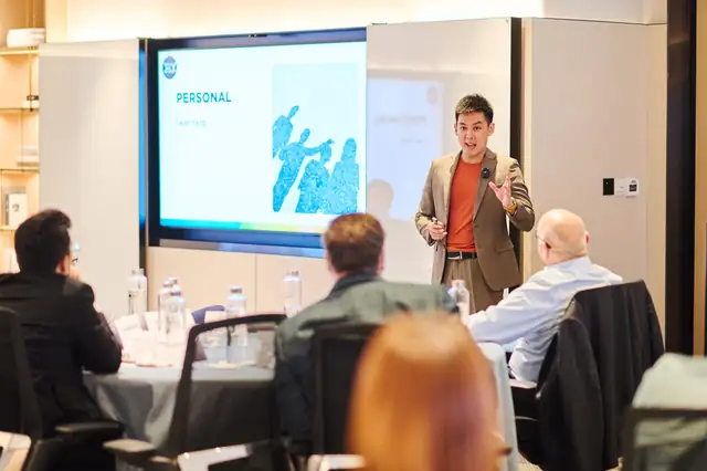 Iwan Yang presenting to a small group around a meeting table, with slides on the screen behind him