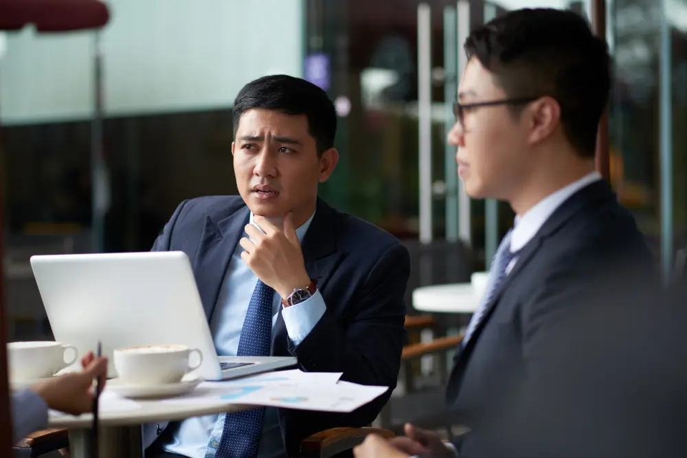 Two colleagues in suits discussing work over a laptop at a cafe table