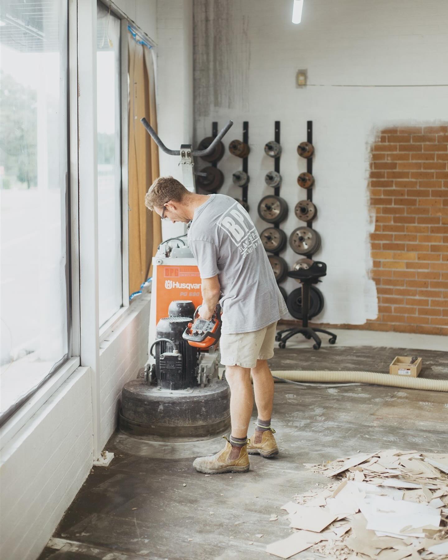 Operator running a floor grinder beside a window, with lifted floor covering stacked on the slab