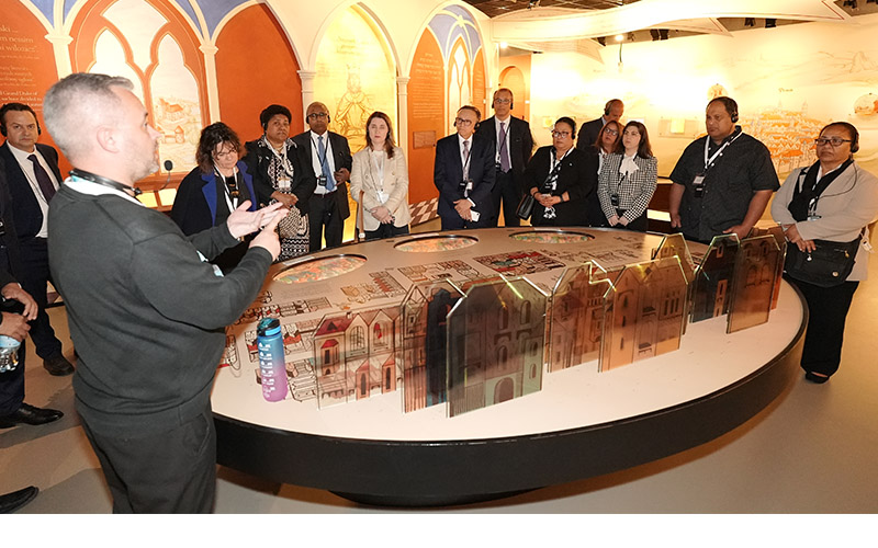 Delegation participants listen to a speaker beside a large display model.