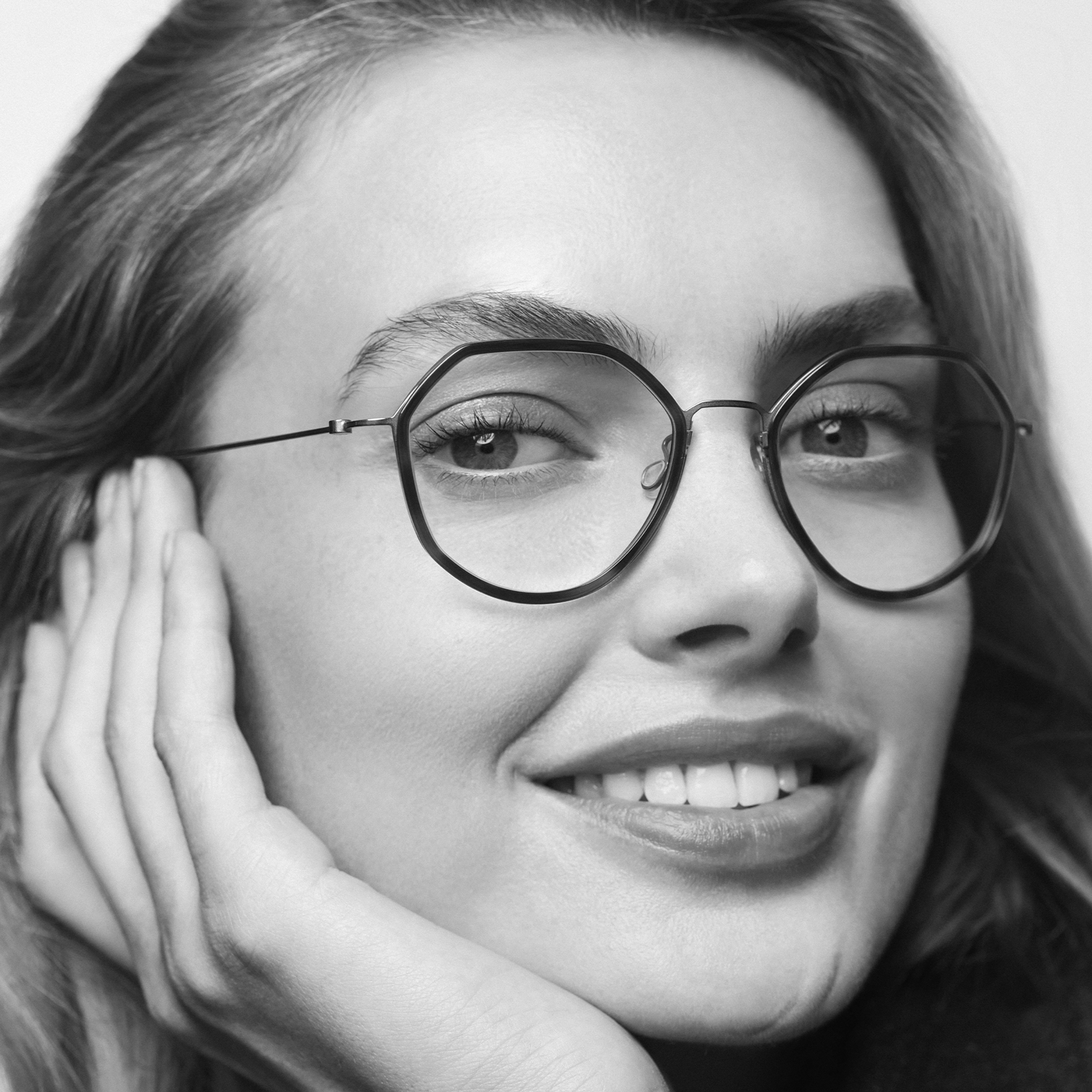 Close-up black and white portrait of a smiling woman with glasses resting her face on her hand.
