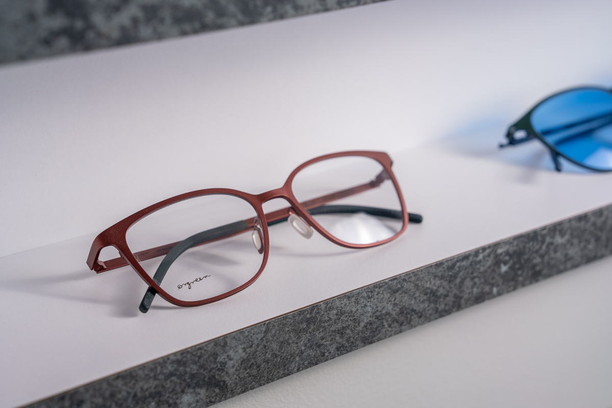 Red-brown rectangular eyeglasses with black inner arms placed on a white surface with a gray edge; a pair of blue-lens sunglasses is partially visible in the background.
