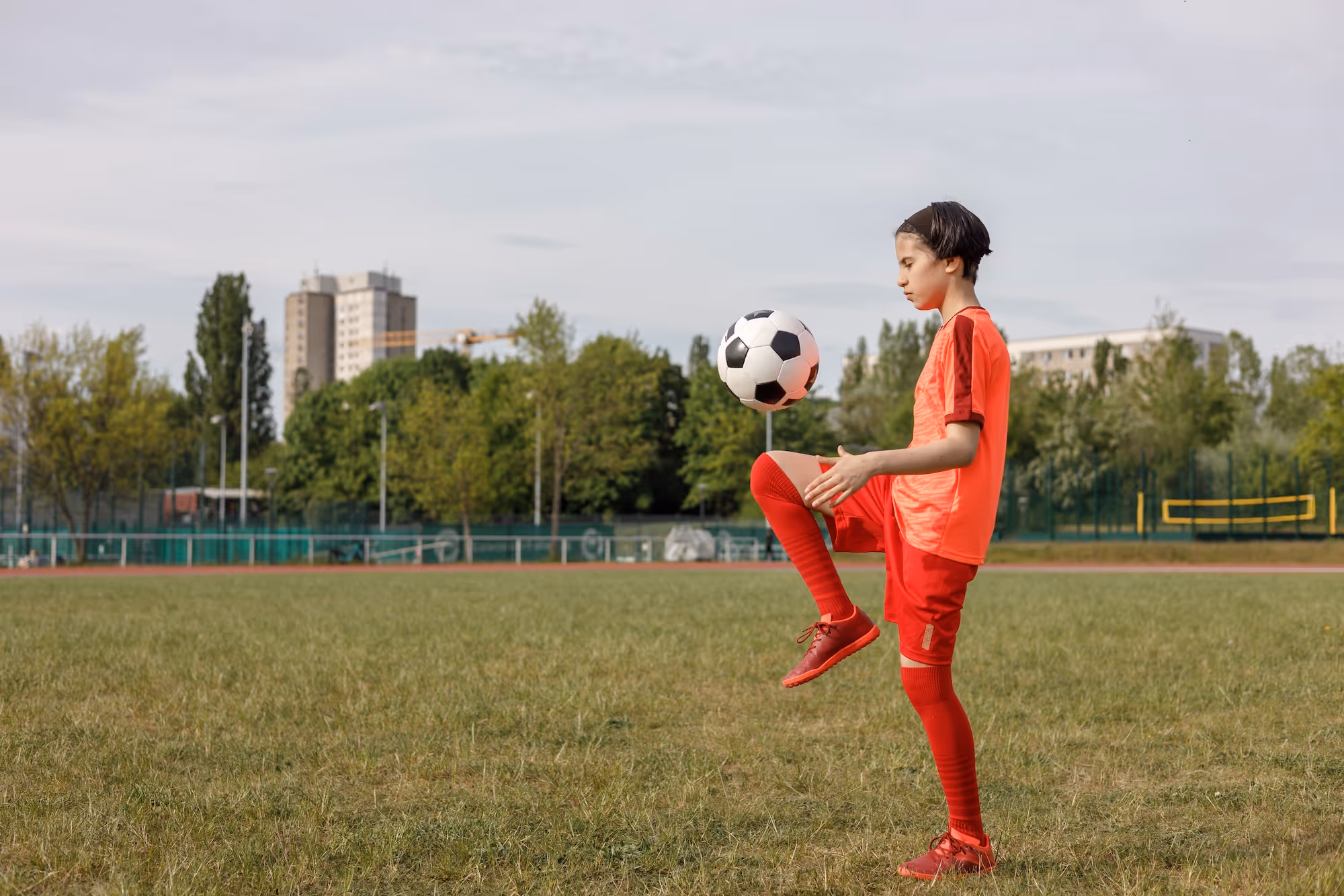 Boy in red sportswear and cleats balancing a black and white soccer ball on his raised right knee on a grassy field with trees and buildings in the background.
