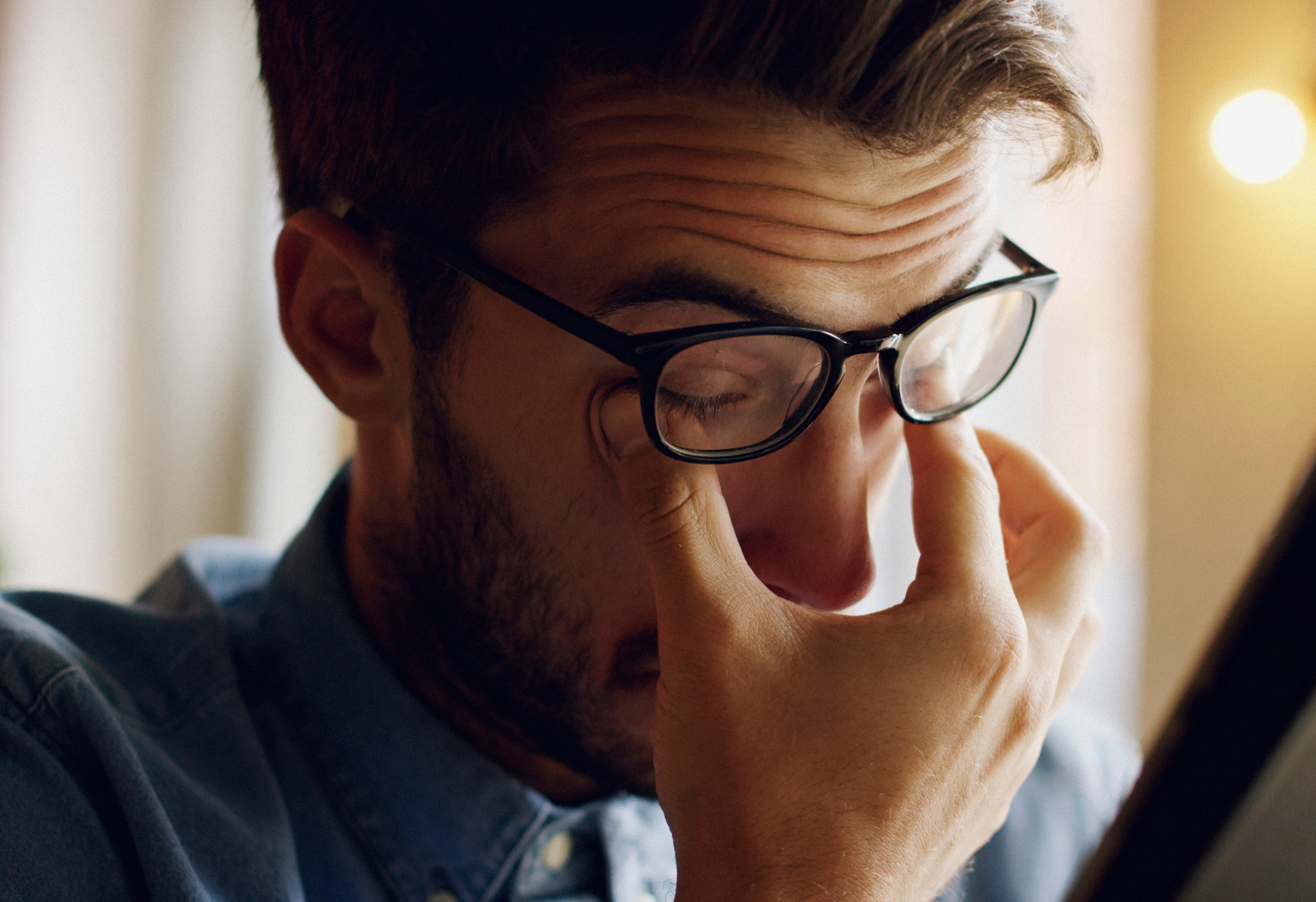 A man with a beard and glasses is pinching the bridge of his nose with his eyes closed, suggesting fatigue or stress.
