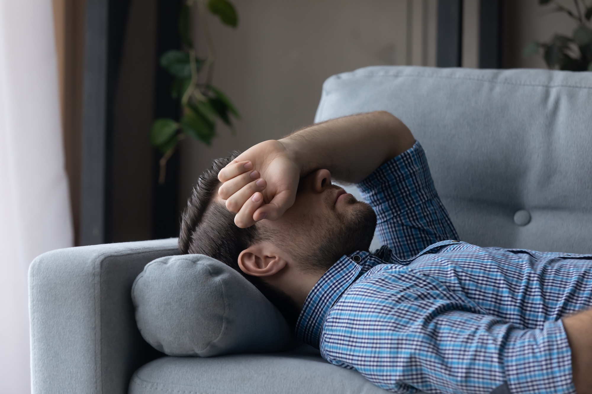 A man in a blue plaid shirt lying on a light gray couch with his head on a pillow and his arm covering his eyes, appearing tired or stressed.