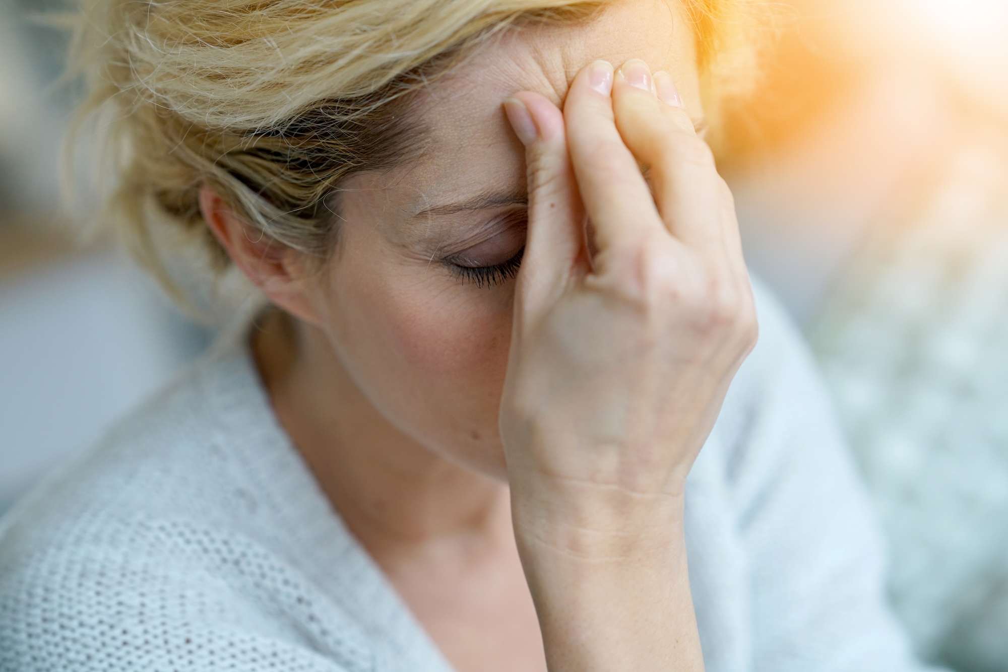 Close-up of a woman with blonde hair holding her forehead with her eyes closed, appearing stressed or in pain.