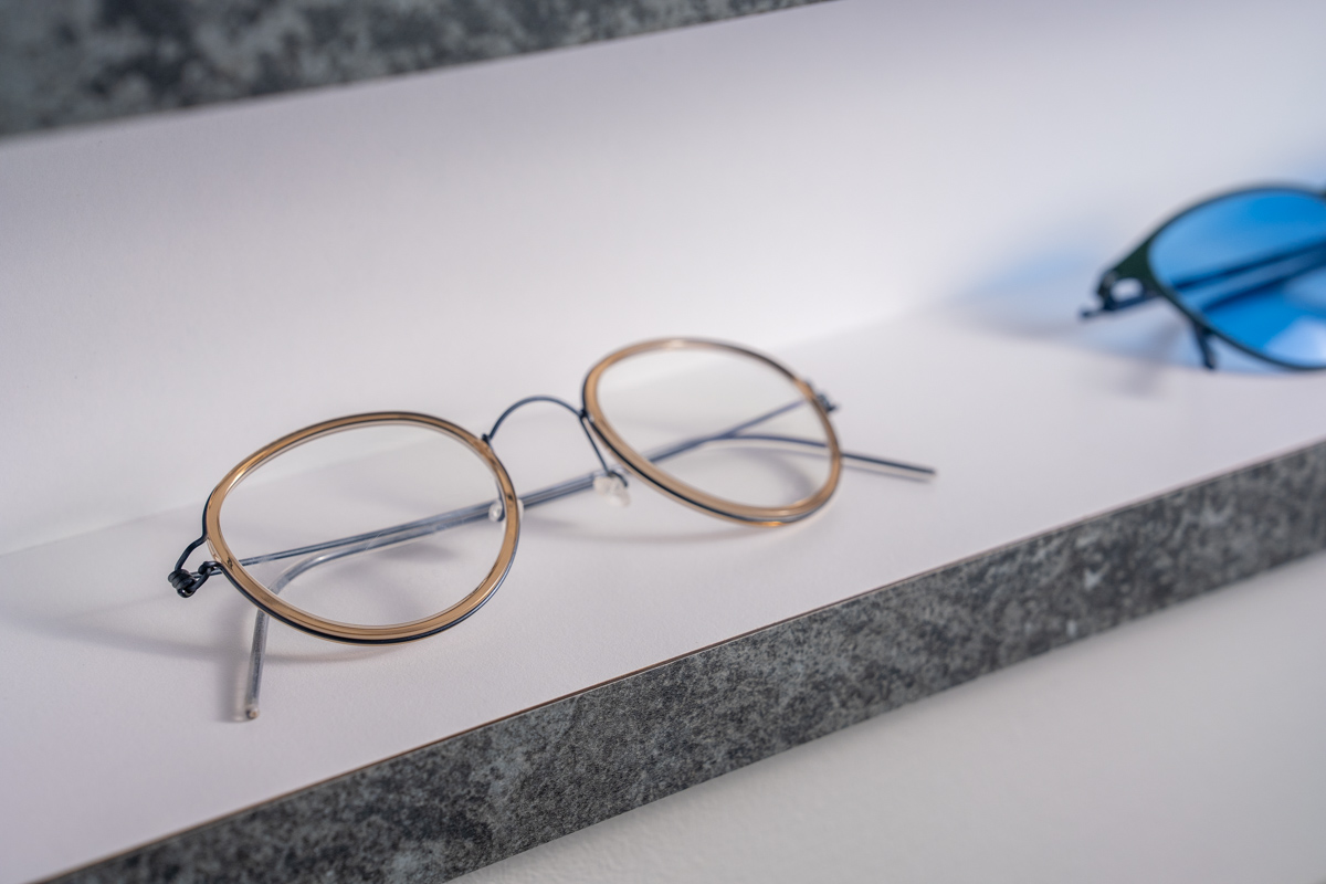 A pair of round eyeglasses with thin gold-rimmed frames and silver temple arms resting on a white surface, with a blurred pair of blue-lensed sunglasses in the background.