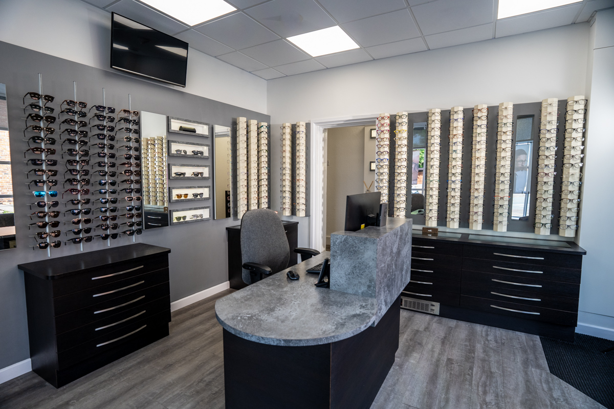 Modern optometry office interior with multiple racks displaying eyeglasses and sunglasses on gray walls, a gray office chair behind a curved gray and black reception desk with computer monitor, keyboard, and mouse, and wood-look flooring.