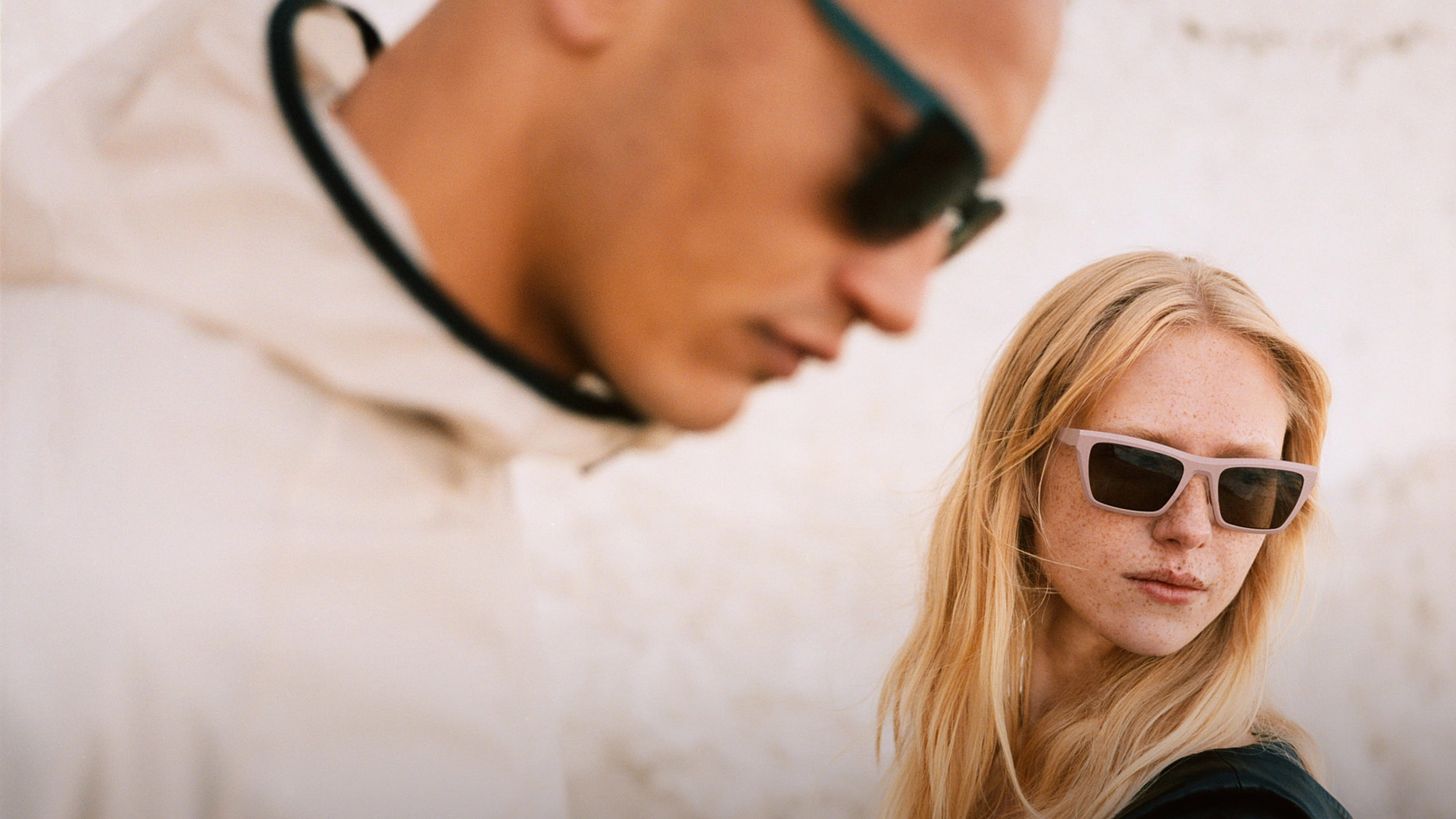 Close-up of a young woman with long blonde hair and freckles wearing light pink sunglasses looking to the side, with a blurred young man in a white jacket and dark sunglasses in the foreground.
