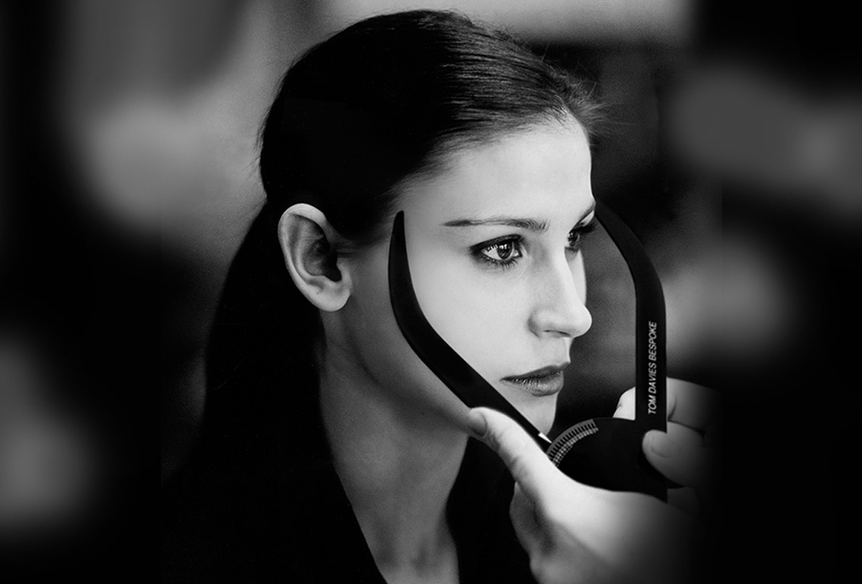 Black-and-white photo of a woman with dark hair pulled back, having her face measured with a device labeled 'Tom Davies Bespoke' held by another person.