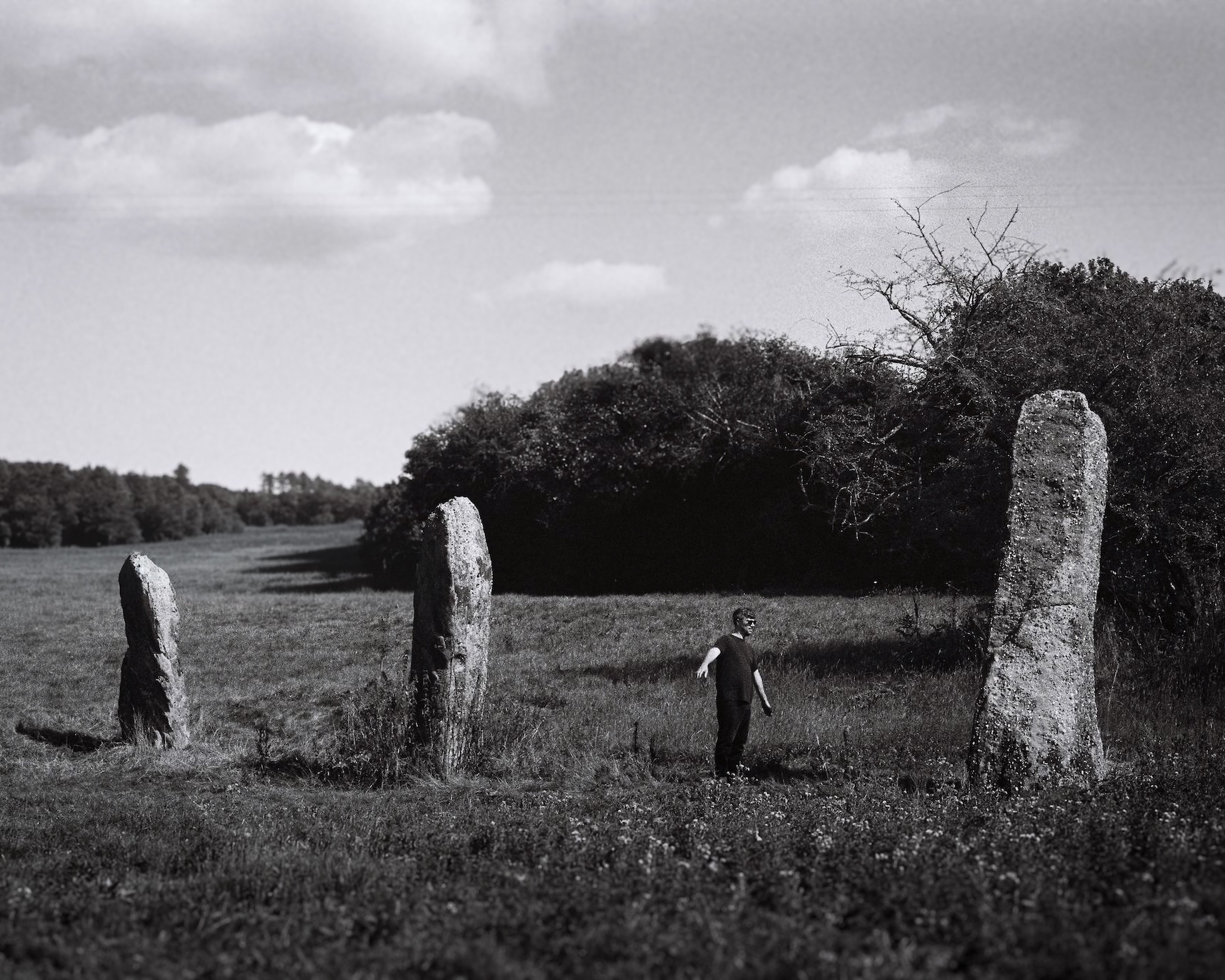 Black and white photo of a man standing in a grassy field near three large, upright standing stones arranged in a rough line with trees and bushes in the background under a partly cloudy sky.