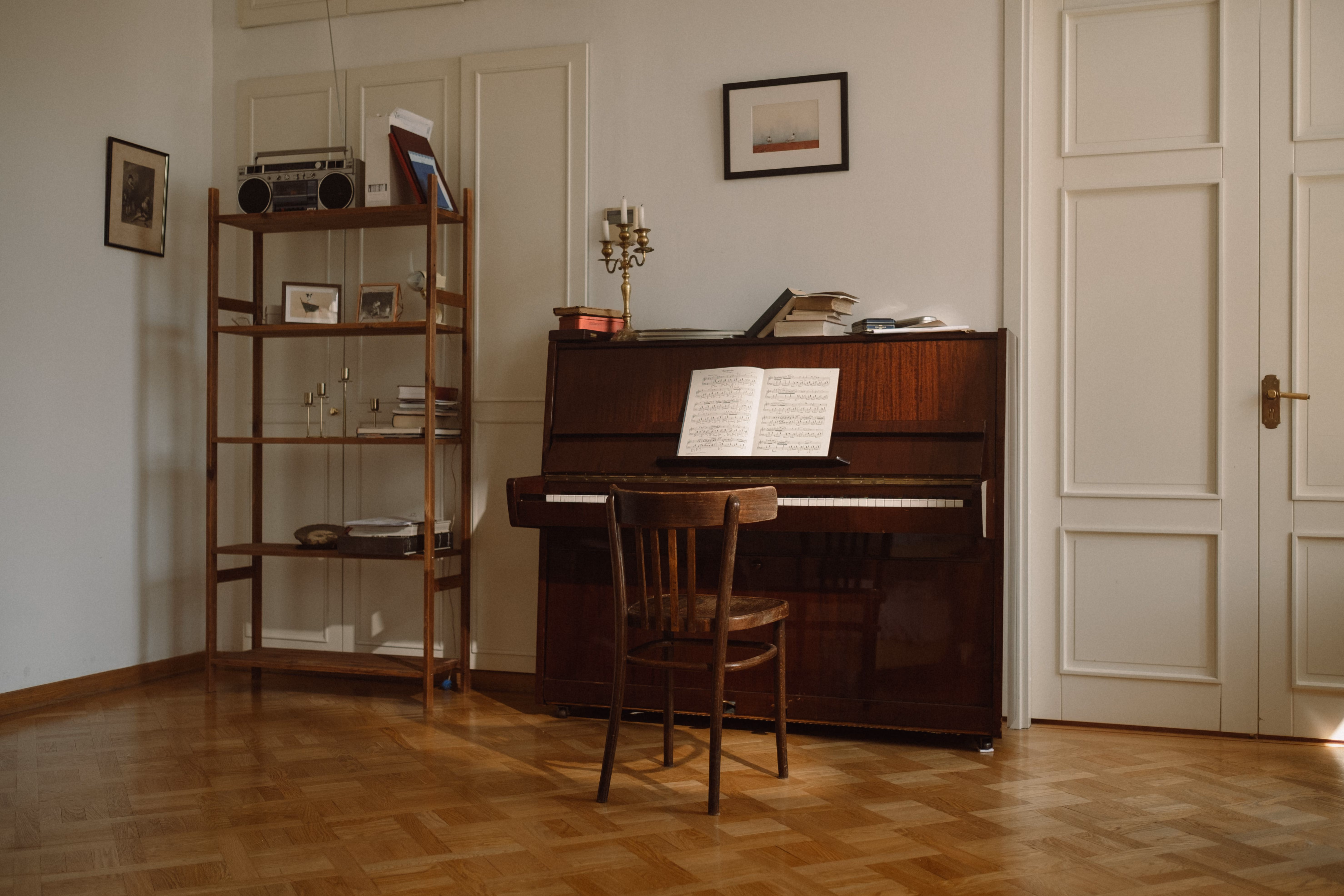 Una stanza con pavimento in parquet chiaro, una sedia di legno scuro davanti a un pianoforte verticale scuro con uno spartito aperto e libri sopra, e a sinistra una libreria con un vecchio registratore e alcuni oggetti decorativi.