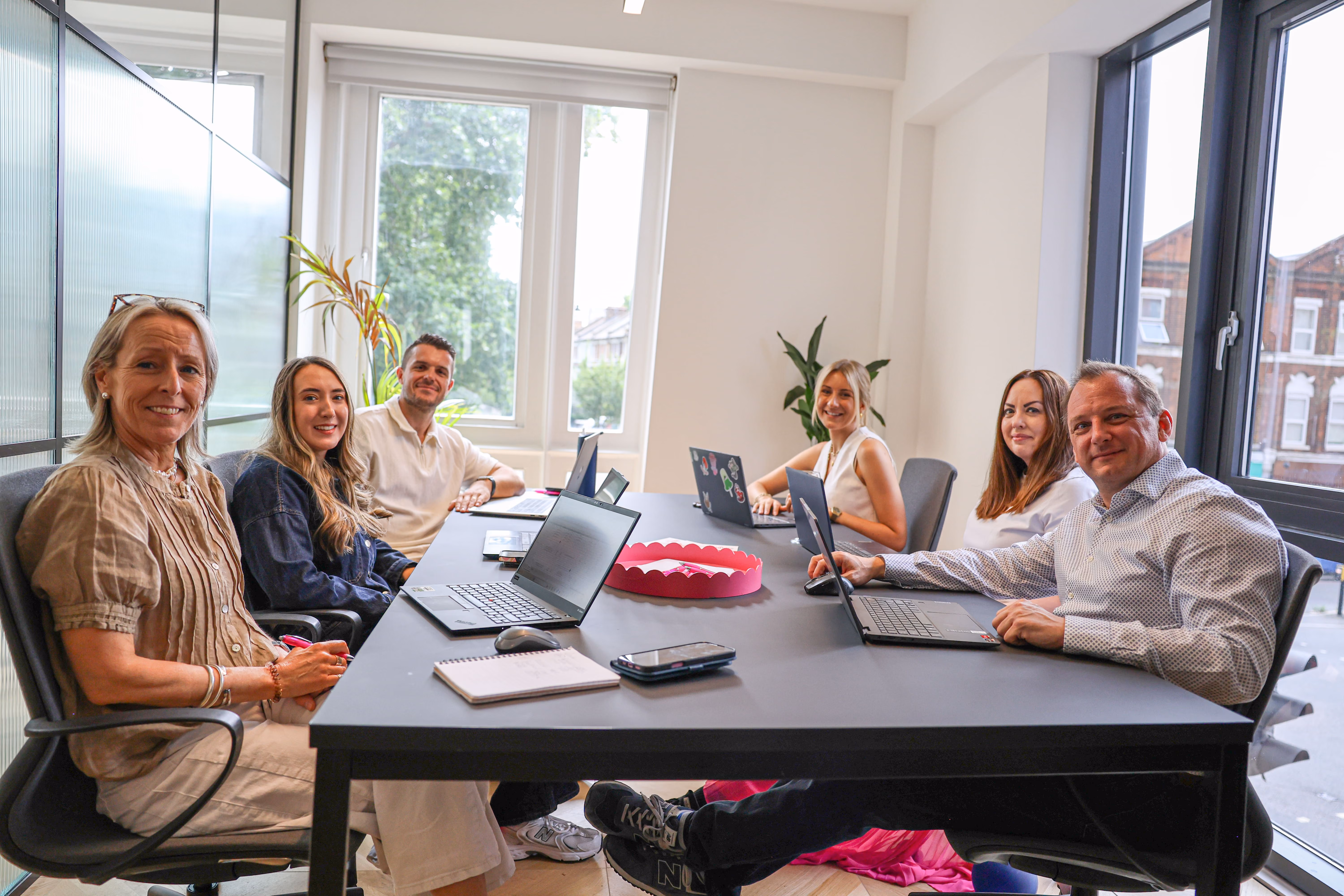 Six people smiling around a conference table in a modern office with laptops and notebooks, large windows letting in natural light.