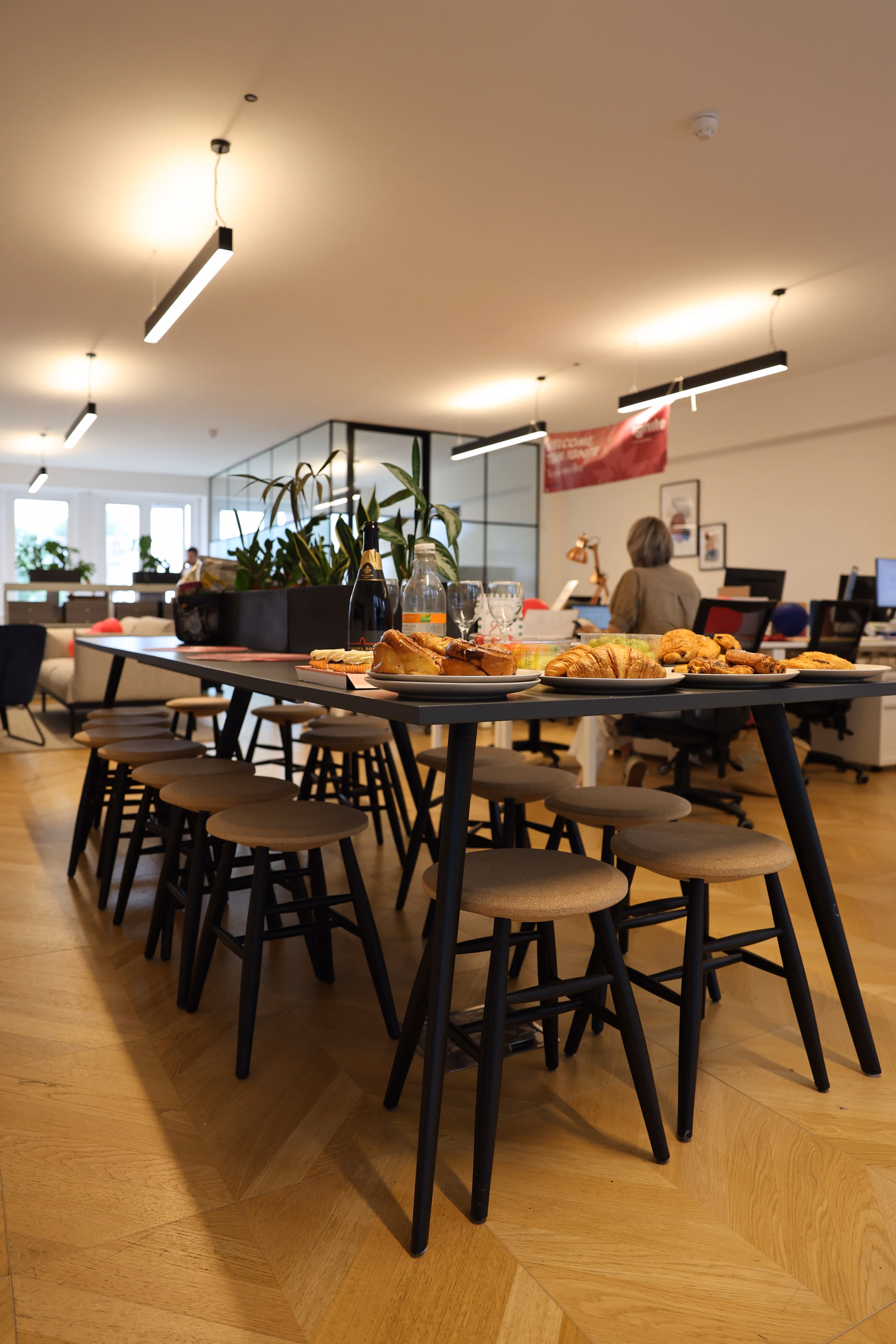 Modern office space featuring a long high table with multiple round stools, topped with plates of croissants and pastries, a bottle, glasses, and plants, with workers and desks in the background.
