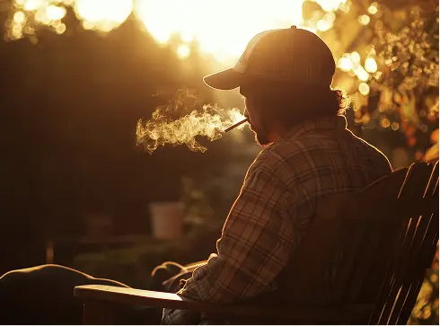 Person wearing a cap and plaid shirt sitting outdoors in a wooden chair, smoking and exhaling smoke illuminated by warm sunlight.