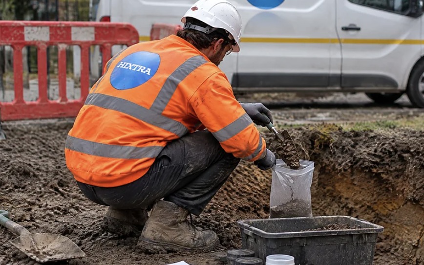 Hixtra technician in hi-vis crouching to take a soil sample from a trial pit