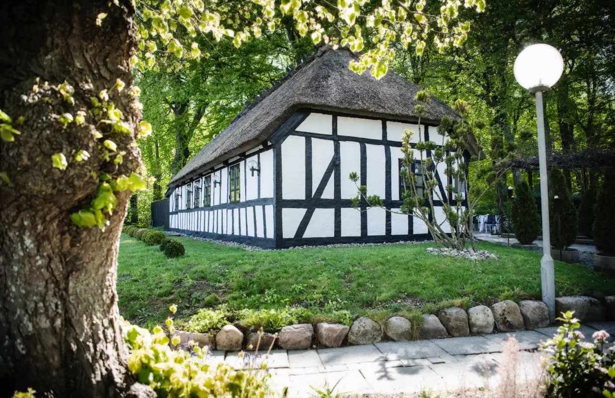 Skovhusets bindingsværksgavl og stråtag omgivet af grønne træer.