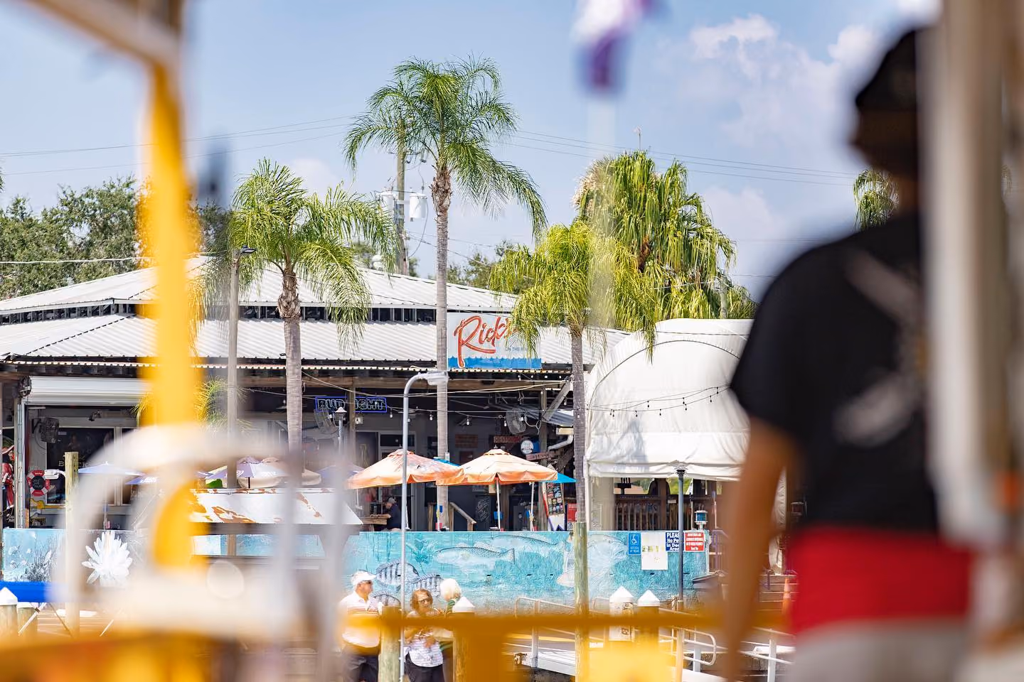 Outdoor waterfront dining area with palm trees, colorful umbrellas, and a building with the sign 'Rick's' in the background, seen through blurred foreground elements including a person in red shorts and a black shirt.