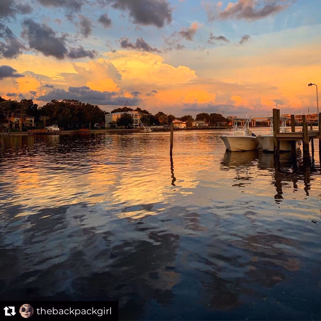 Calm waterway at sunset reflecting orange, yellow, and blue sky clouds with boats docked near a wooden pier and houses along the shoreline.
