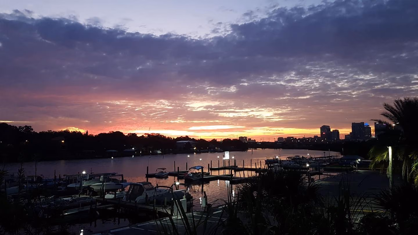 Sunset over a calm river with docks and boats in the foreground, city skyline and trees in the background, and a cloudy sky illuminated with purple, pink, and orange hues.