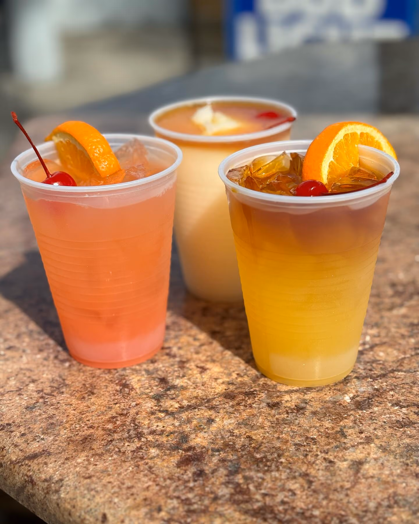 Three plastic cups on a stone surface filled with different cold drinks; the left one is pink with ice, a cherry, and an orange slice, the middle is creamy with a cherry, and the right is yellow-orange with ice, a cherry, and an orange slice.