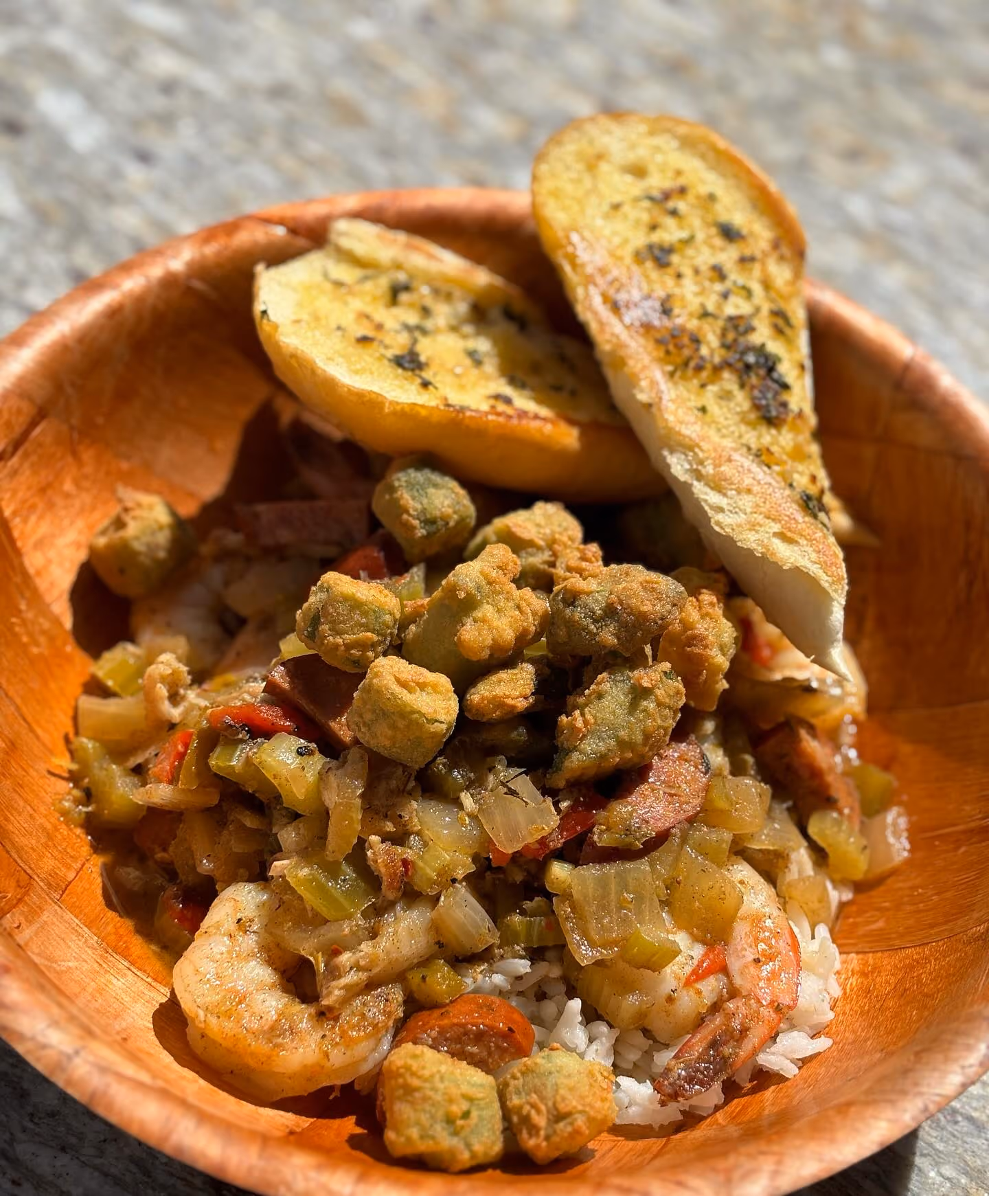 Wooden bowl containing a Cajun-style shrimp and sausage rice dish topped with fried okra and served with two slices of garlic bread.