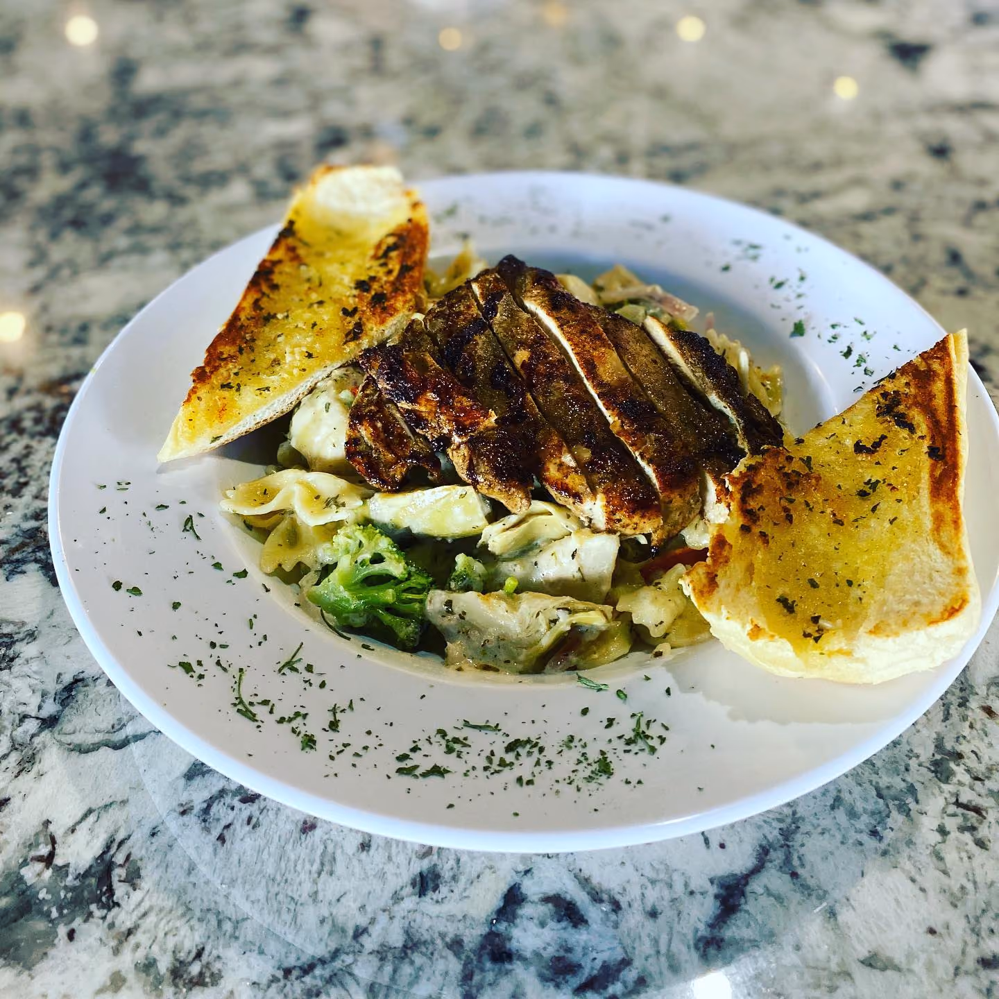 White plate with grilled chicken slices over creamy pasta containing bow ties and broccoli, garnished with herbs around the rim and served with two pieces of toasted garlic bread on a marble surface.