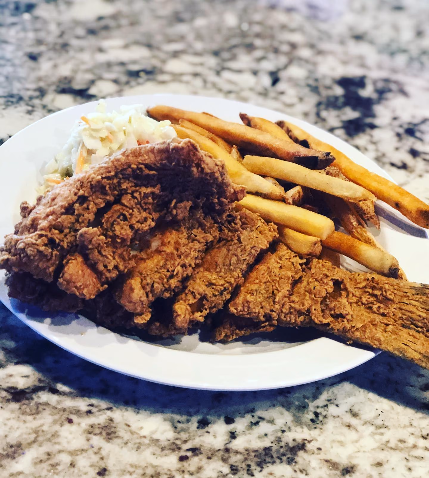 White plate with fried catfish filet, golden French fries, and coleslaw on a speckled granite countertop.