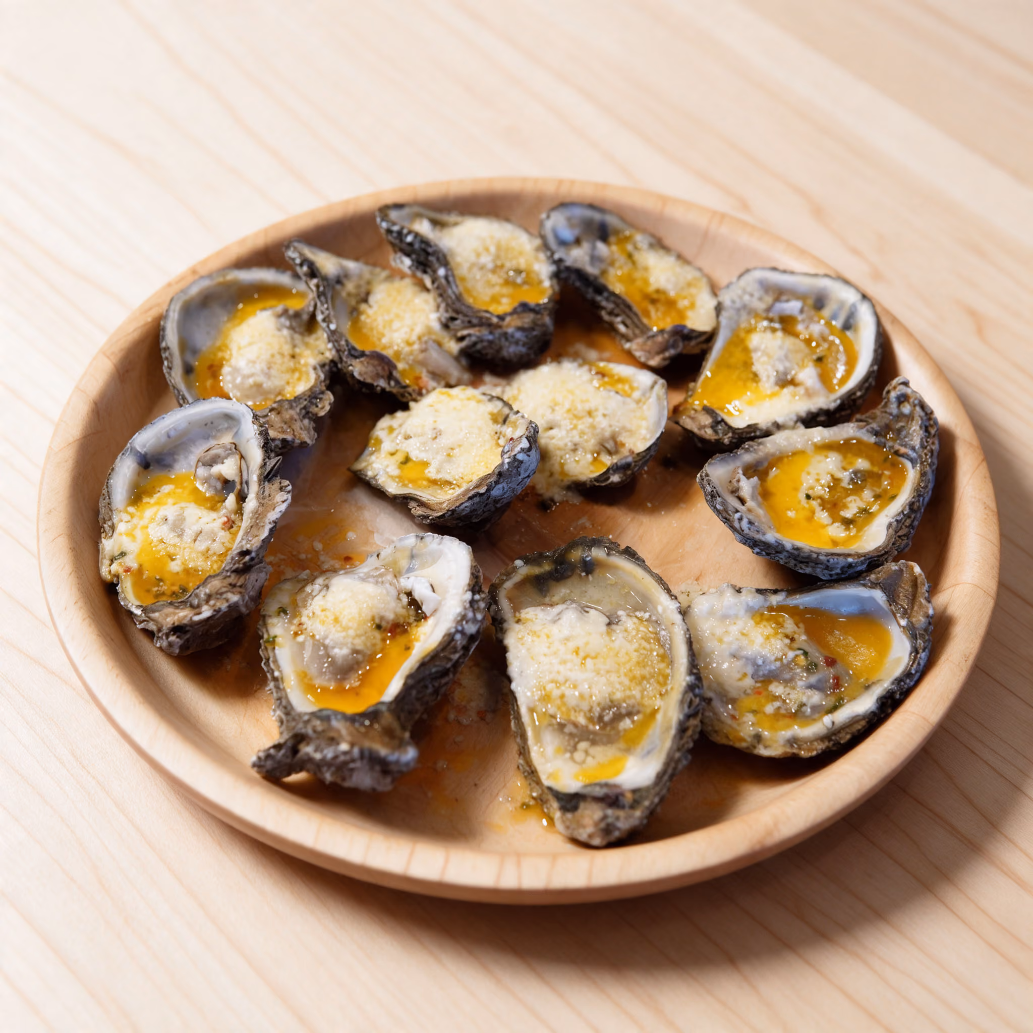 A circular wooden plate holding twelve baked oyster shells topped with melted butter, herbs, and grated cheese, placed on a light wooden surface.