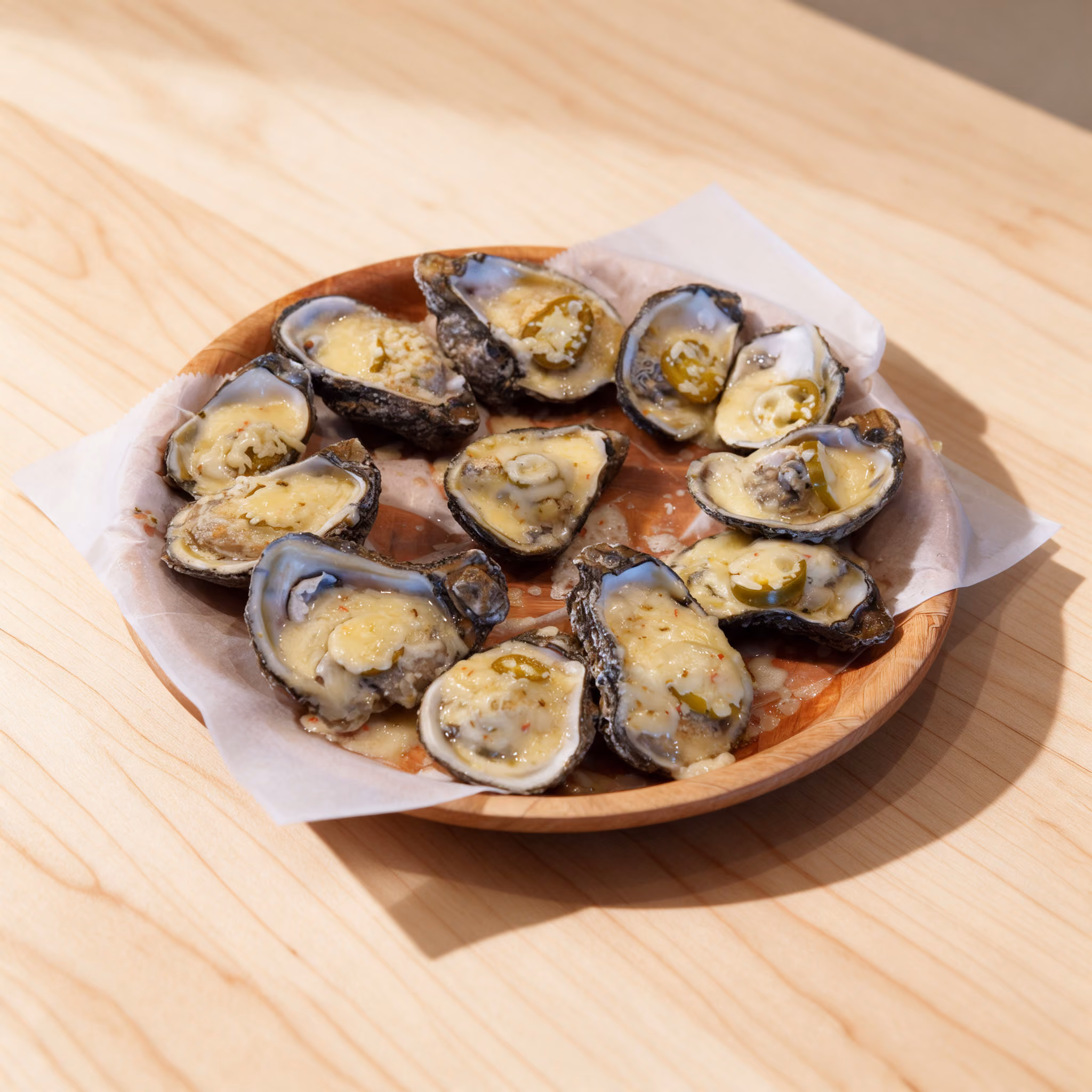 Wooden plate with multiple oyster shells topped with melted cheese and slices of jalapeño on parchment paper, placed on a light wood surface.