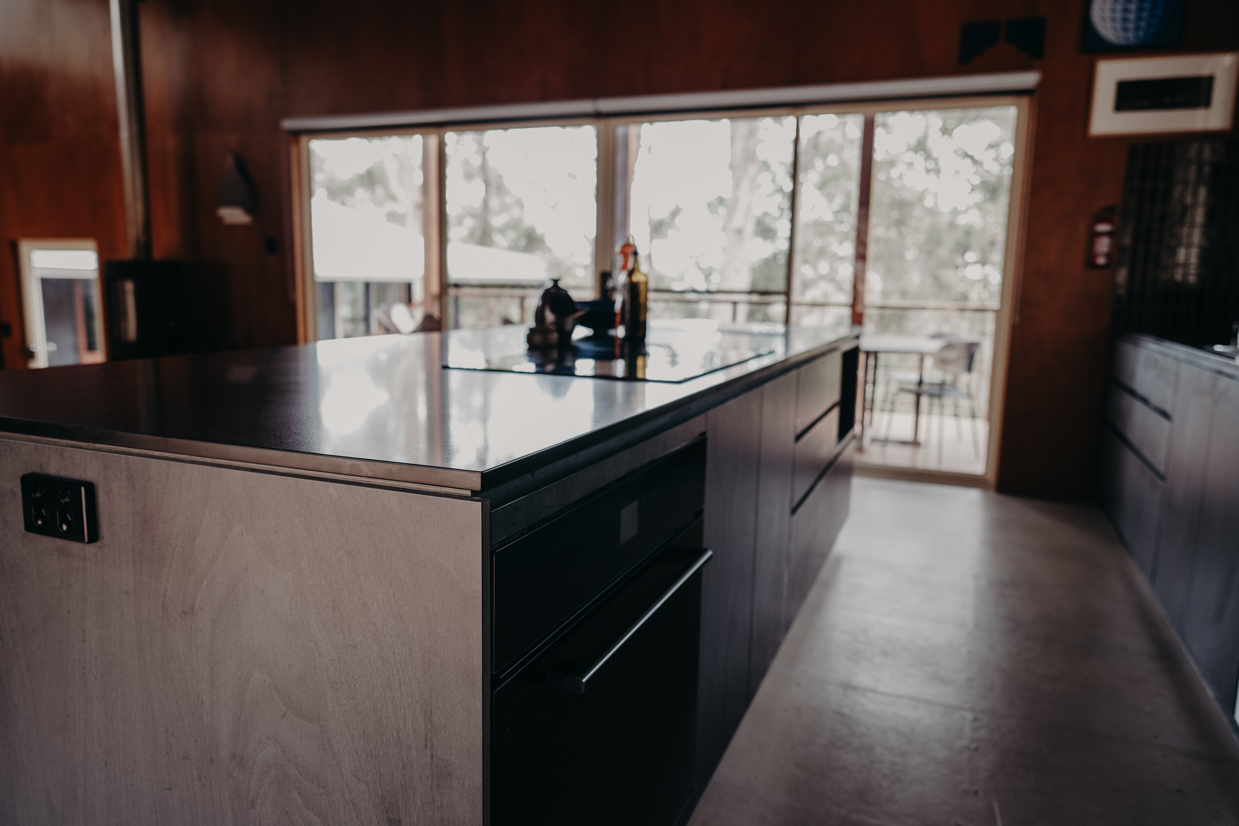 Kitchen island with a stainless benchtop and dark joinery looking out to the deck of a renovated Denmark home