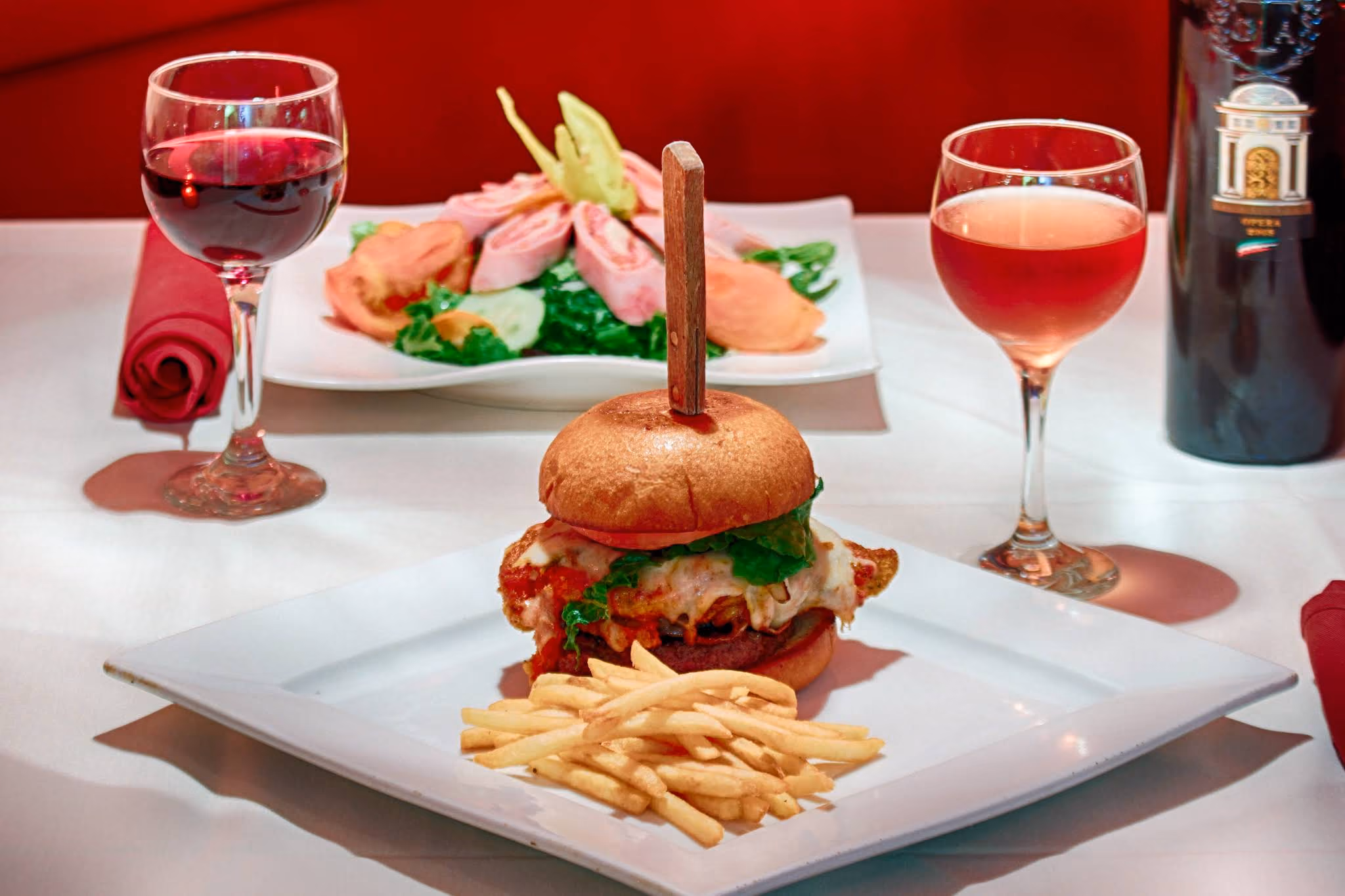 Grilled chicken sandwich with melted cheese and lettuce served with French fries on a square white plate, accompanied by a glass of red wine and a glass of rosé wine on a white table with a red backdrop.