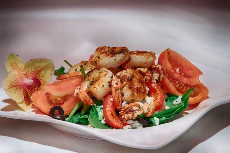 Grilled shrimp served on a bed of spinach leaves with sliced tomatoes, black olives, and crumbled cheese, garnished with a yellow orchid flower on a white plate.