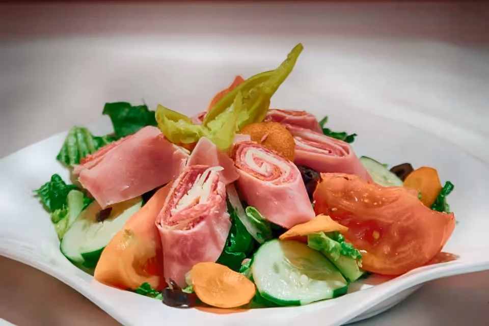 Salad with rolled ham slices, fresh tomato wedges, cucumber slices, carrot rounds, leafy greens, and a pepper garnish arranged on a white plate.
