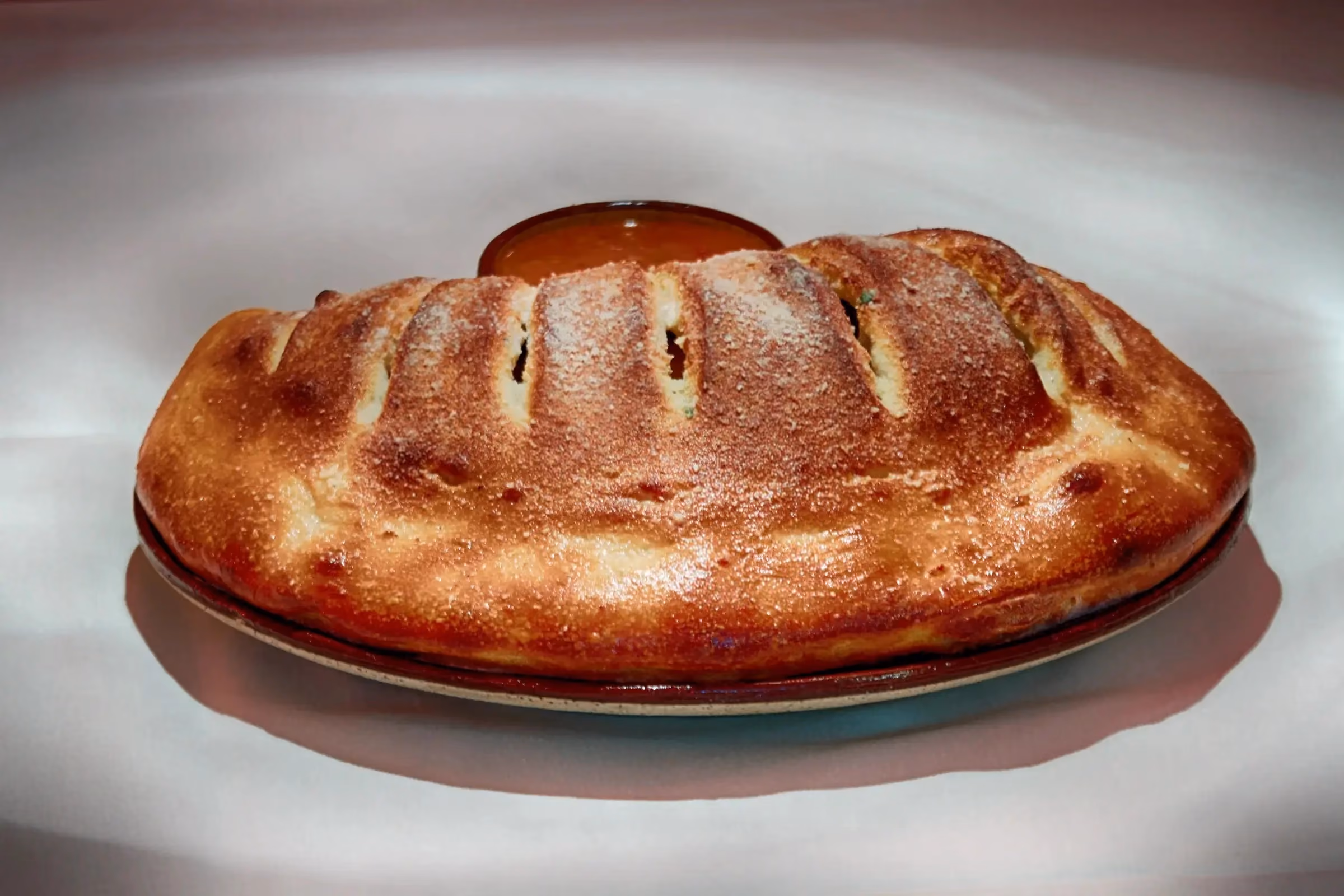Golden-brown baked calzone with slits on top, served on a round plate with a small bowl of red dipping sauce behind it on a white surface.