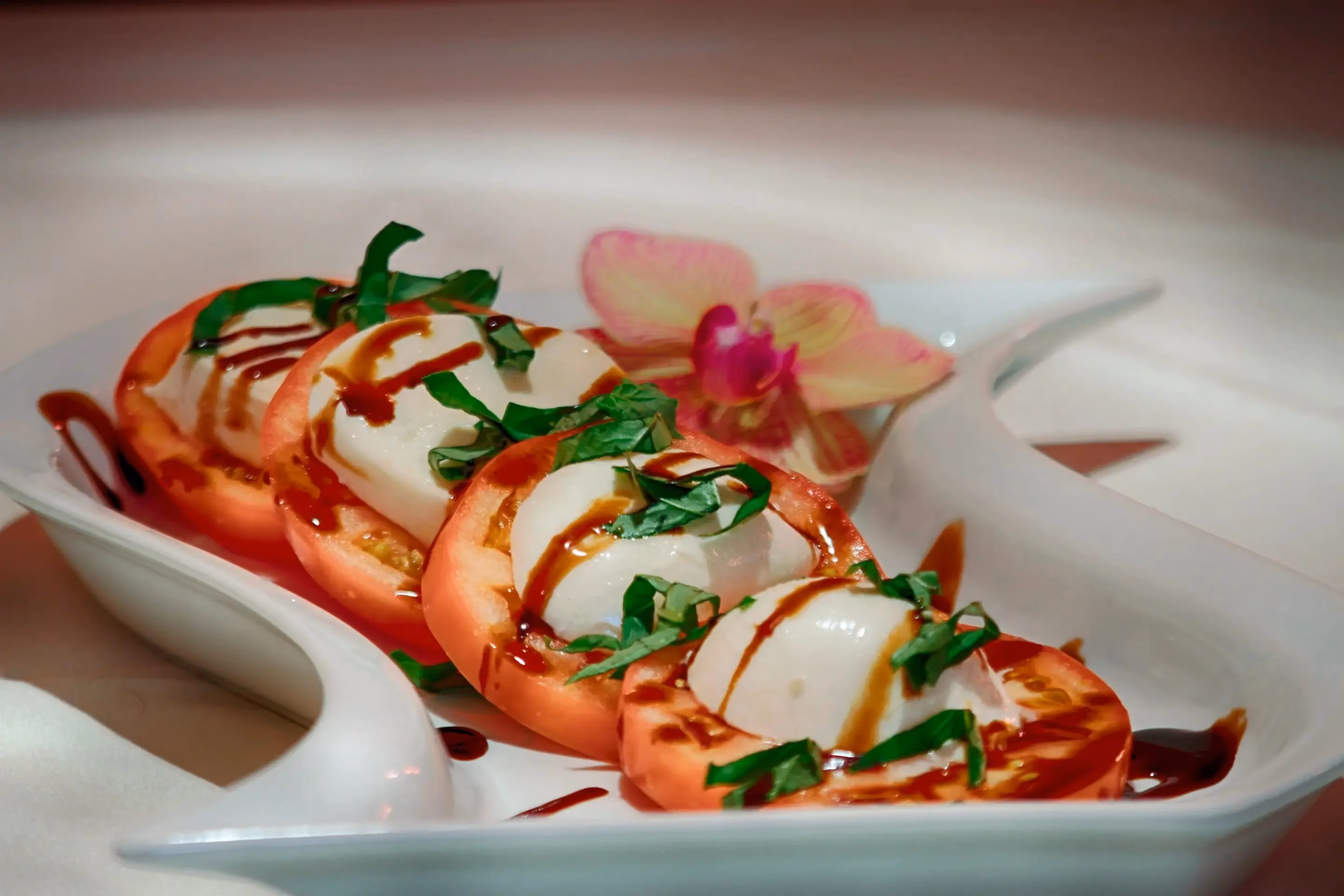 Caprese salad with sliced tomatoes and mozzarella cheese topped with chopped basil and drizzled balsamic glaze served on a white plate with a pink orchid flower garnish.