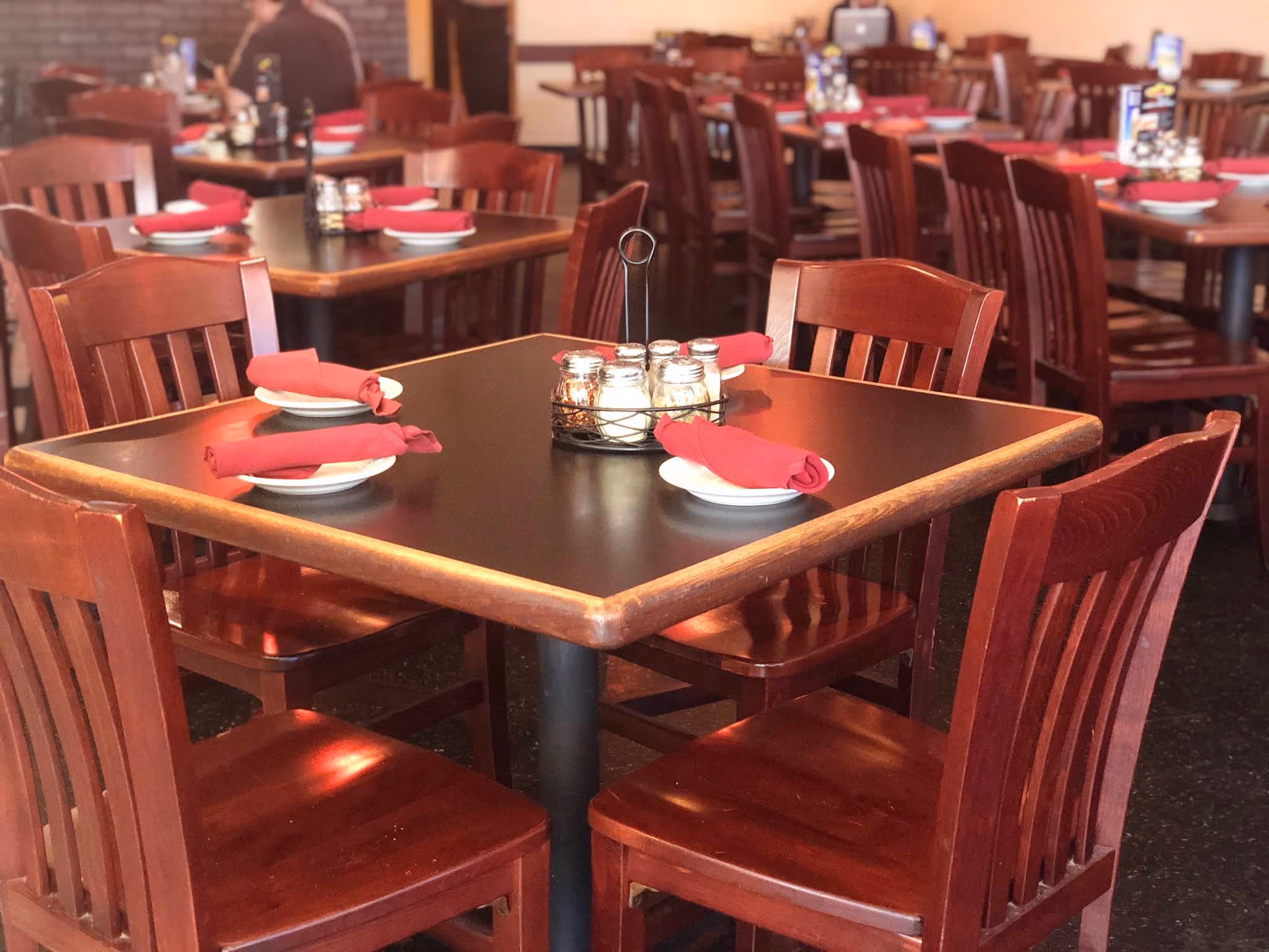 Restaurant interior with wooden tables and chairs, tables set with white plates and red cloth napkins, and condiment caddies in the center.