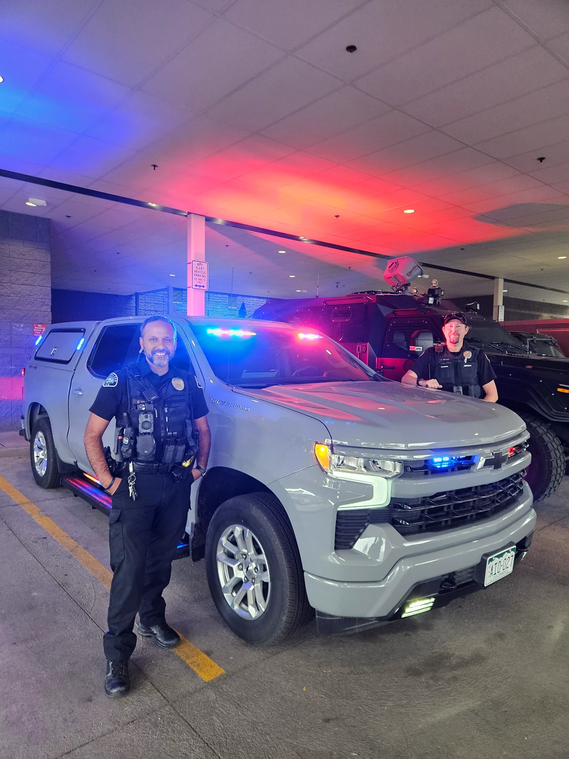 Two uniformed police officers stand beside a gray police pickup truck with its red and blue emergency lights on in a parking garage.