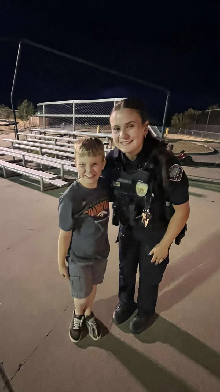 A smiling uniformed police officer poses with a young boy at night beside the bleachers of a ball field.