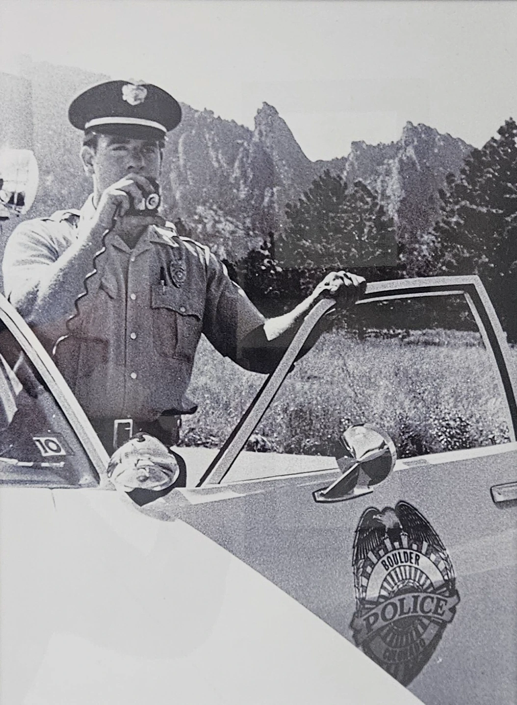 Historical black-and-white photo of a Boulder police officer using a radio beside his patrol car with the Flatirons behind him.