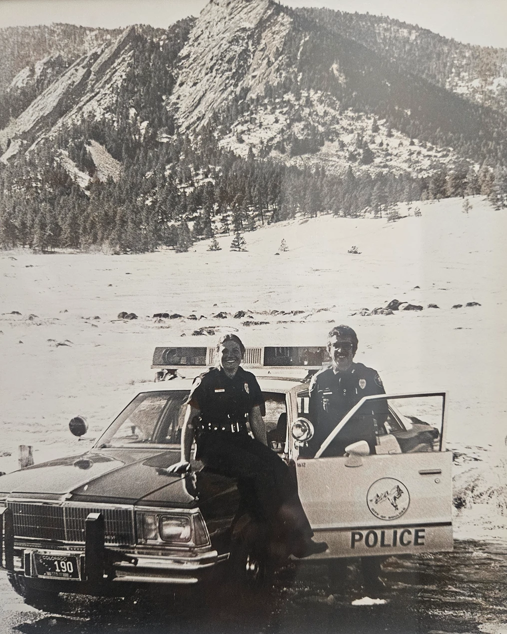 Historical black-and-white photo of two Boulder police officers posing with their patrol car in the snow beneath the Flatirons.